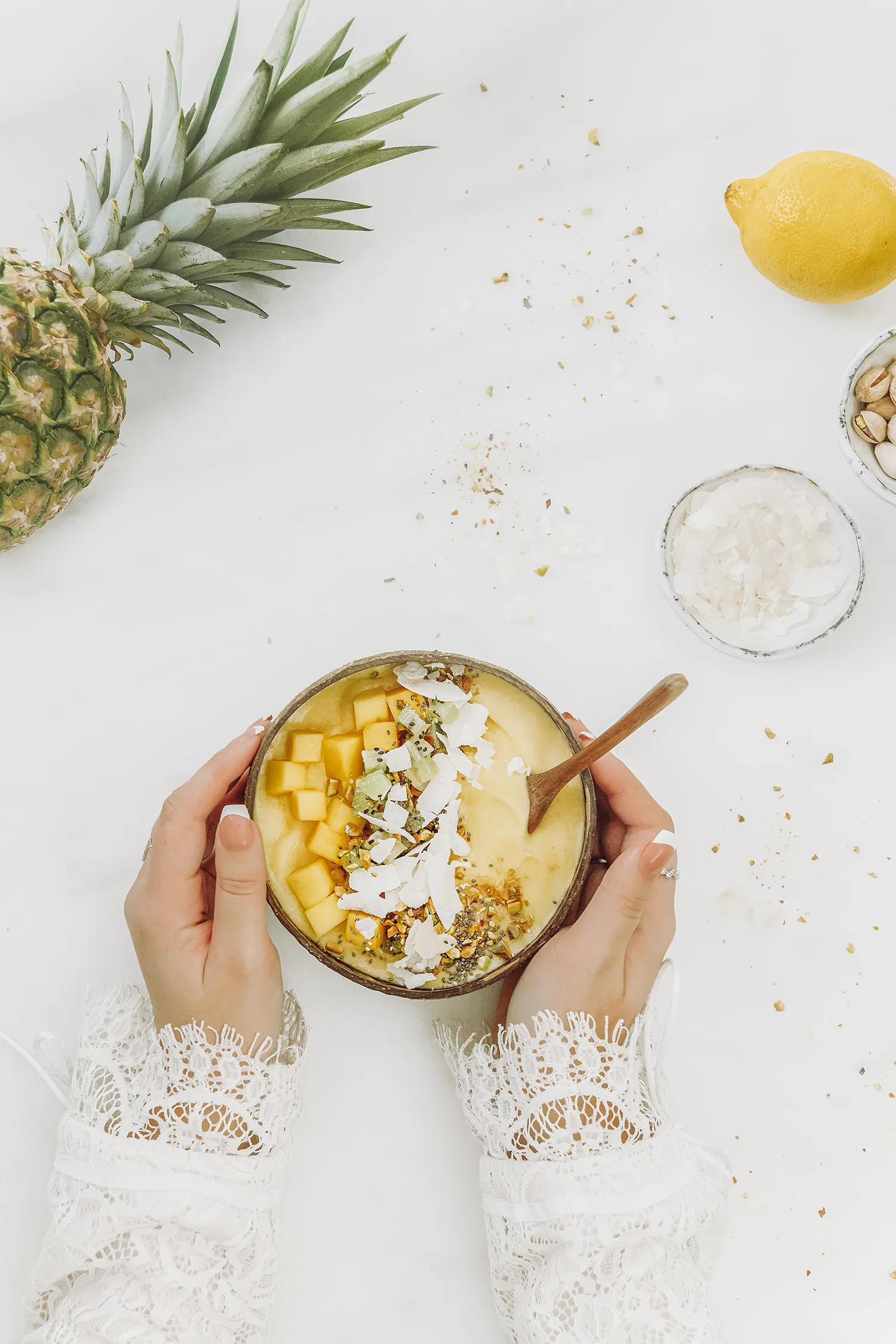 Overhead view of a mango and pineapple smoothie bowl topped with fresh mango, coconut flakes and granola, held in hands on a light background with pineapple nearby.