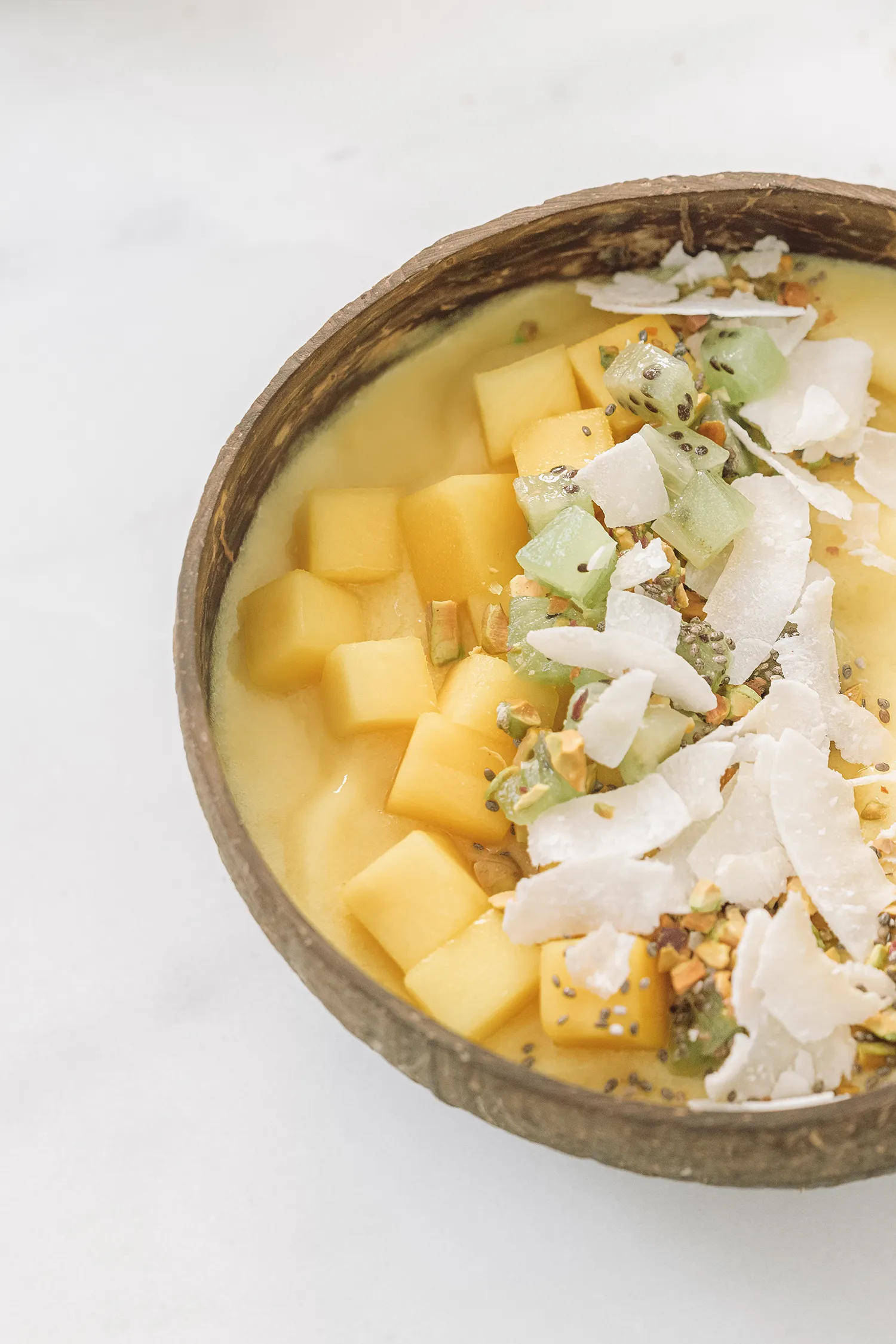 Creamy mango and pineapple smoothie bowl topped with diced fresh mango, kiwi dices, coconut flakes and chia seeds on a light background.
