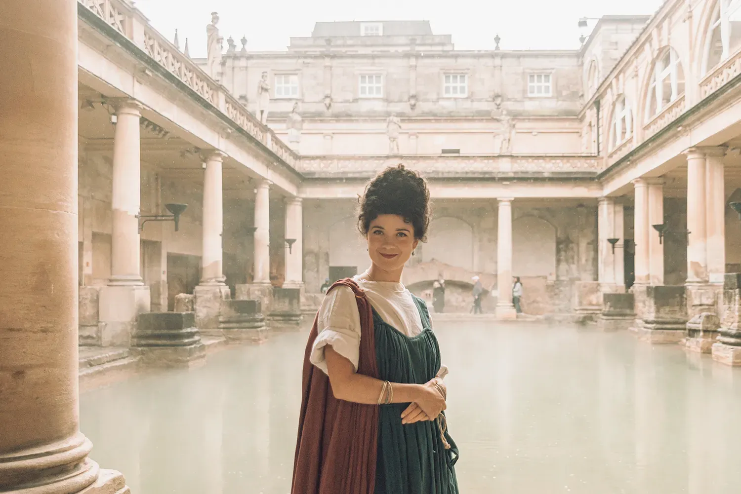 Woman dressed in historical costume at the Roman Baths in Bath, England, with the steaming Great Bath in the background.
