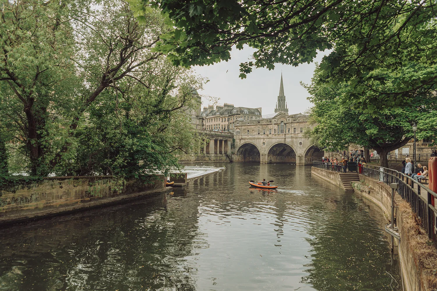 Pulteney Bridge over the River Avon in Bath, England, with kayakers on the water and trees framing the historic bridge.