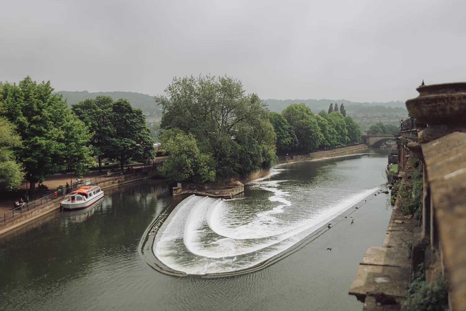 Pulteney Weir on the River Avon in Bath, England, with the curved cascade waterfall and trees along the riverside.