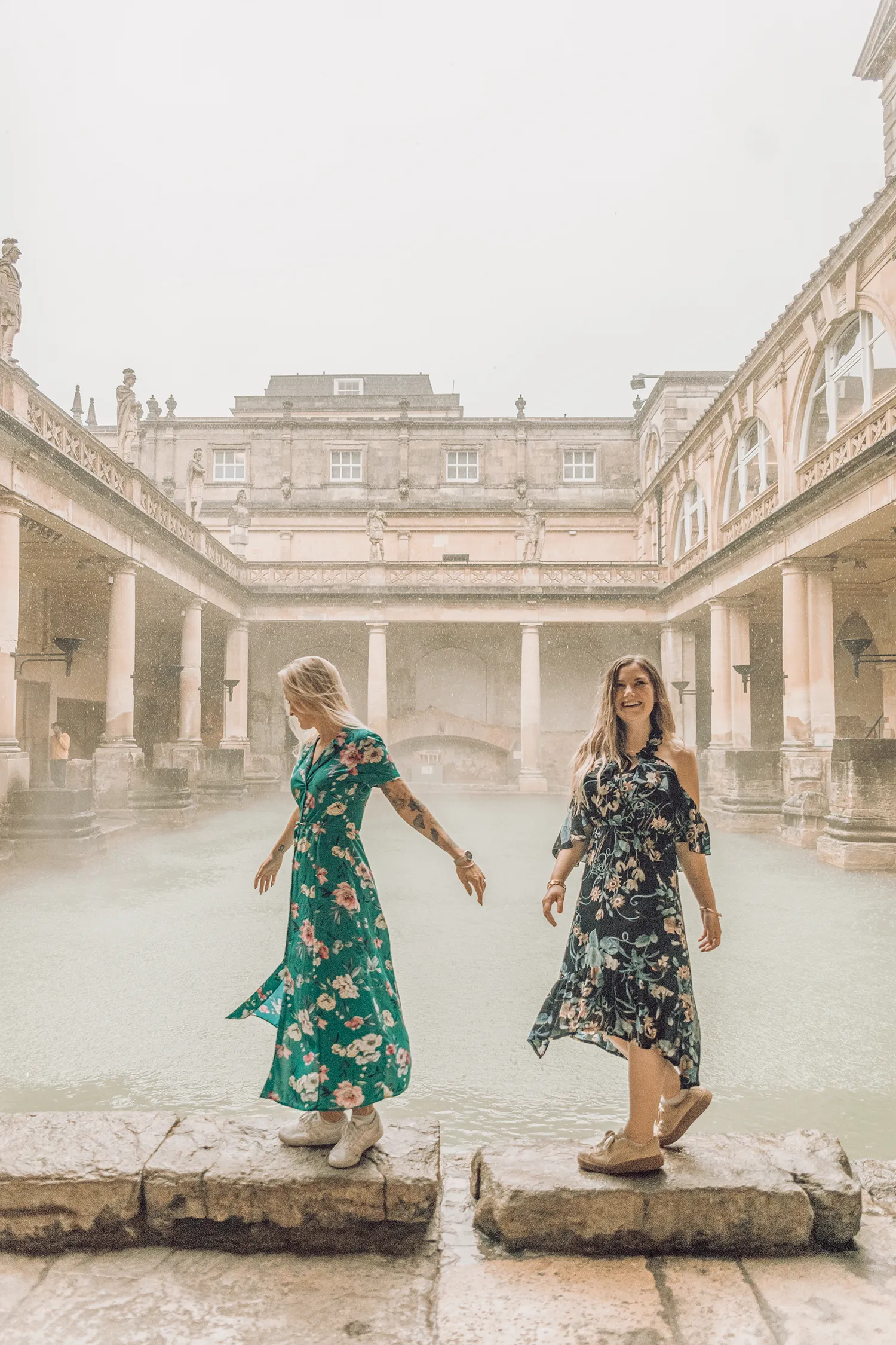 Travel bloggers walking beside the steaming Great Bath at the Roman Baths in Bath, England.