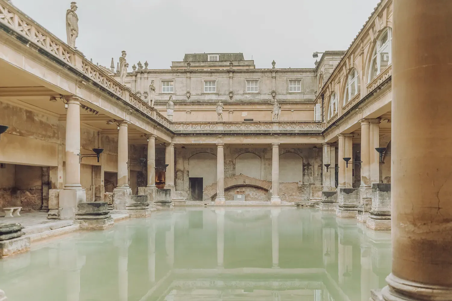 The historic Great Bath at the Roman Baths in Bath, England, with steaming green thermal water and ancient stone columns.